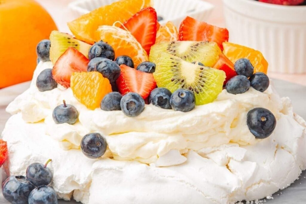 Pavlova dessert topped with whipped cream, blueberries, kiwi, strawberries, and mandarin orange slices.