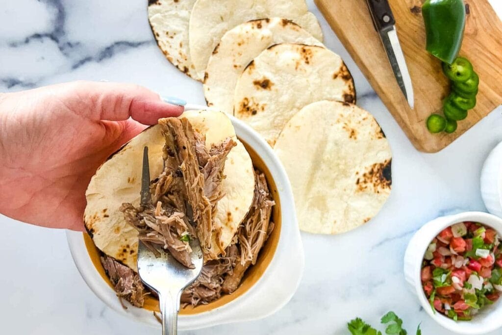 Hand filling a tortilla with shredded pork carnitas.
