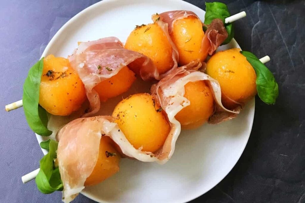 Prosciutto, melon, and basil skewers on a plate.