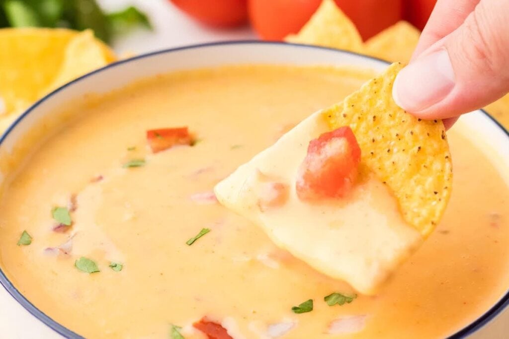 Tortilla chip dipped into creamy queso with tomatoes and cilantro.