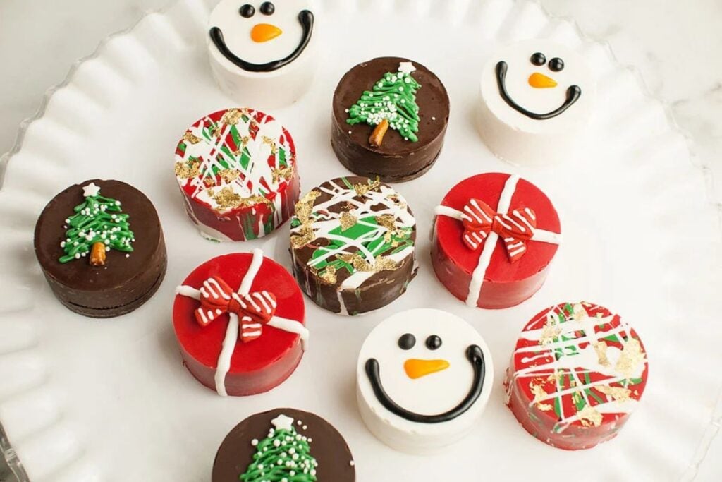 Christmas-themed chocolate-covered Oreos with snowmen, gifts, and Christmas tree decorations on a white plate.