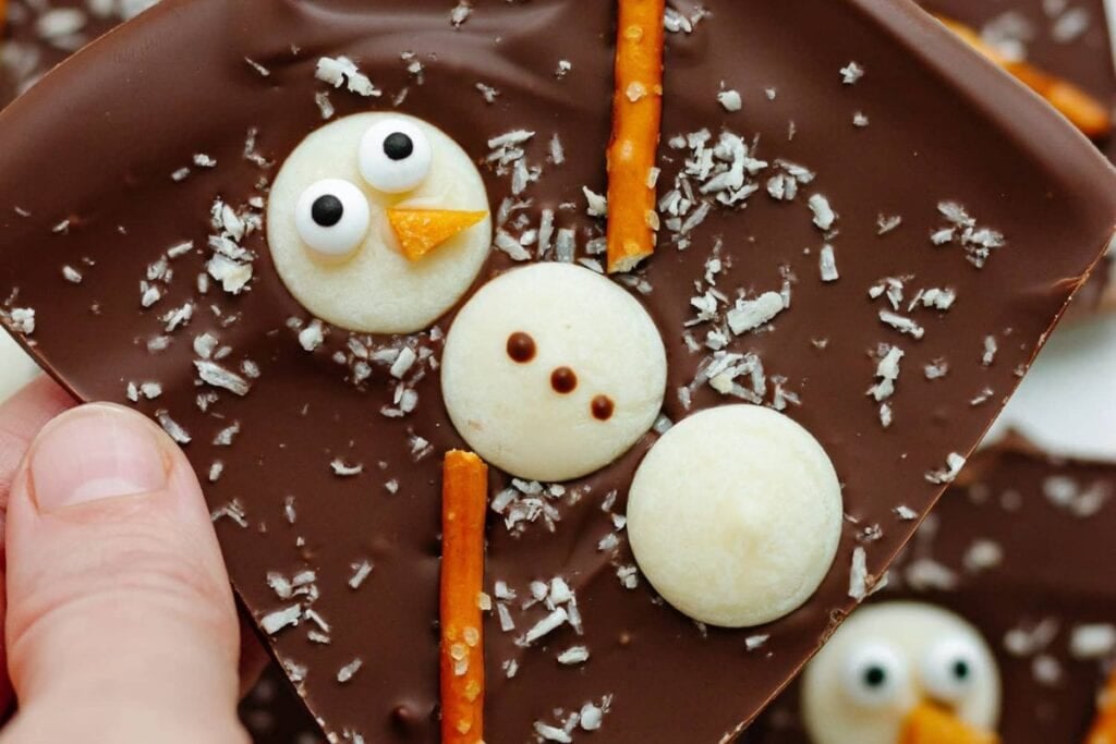 Chocolate bark with candy snowmen, pretzel sticks, and coconut flakes, held by a hand.