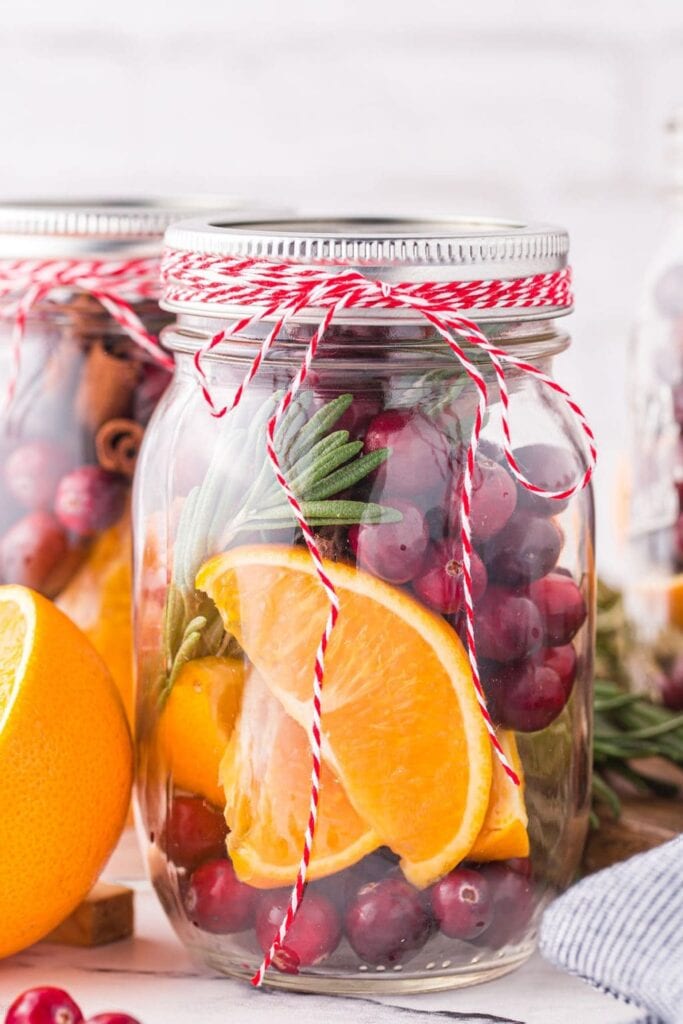 Glass jars filled with orange slices, cranberries, cinnamon sticks, and rosemary sprigs, tied with red-and-white twine and arranged with fresh fruit on a light surface.