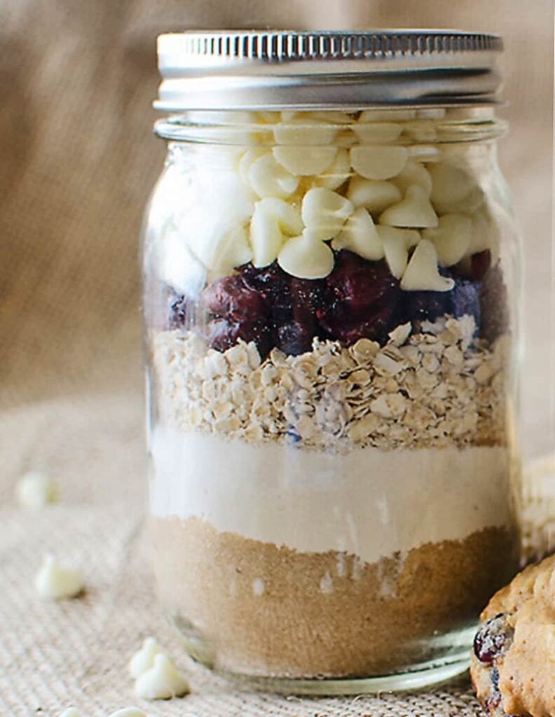 Glass jar layered with brown sugar, flour, oats, dried cranberries, and white chocolate chips, placed on burlap beside a cookie.