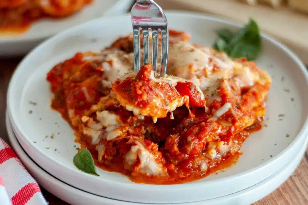 Lasagna slice with tomato sauce and cheese on a white plate with a fork.