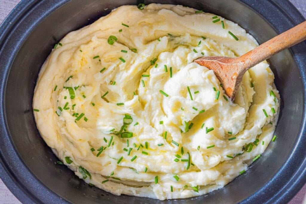 Overhead shot of mashed potatoes with chives in a crockpot with a wooden spoon.
