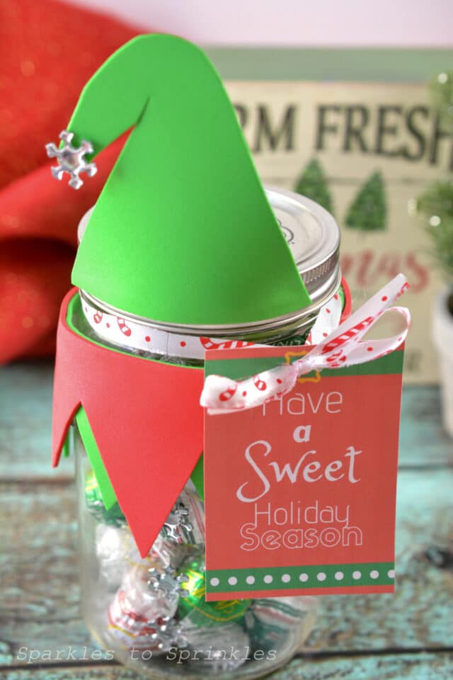 Mason jar decorated as an elf with green hat, red collar, and candy cane ribbon, filled with foil-wrapped candies and festive tag on a rustic surface.