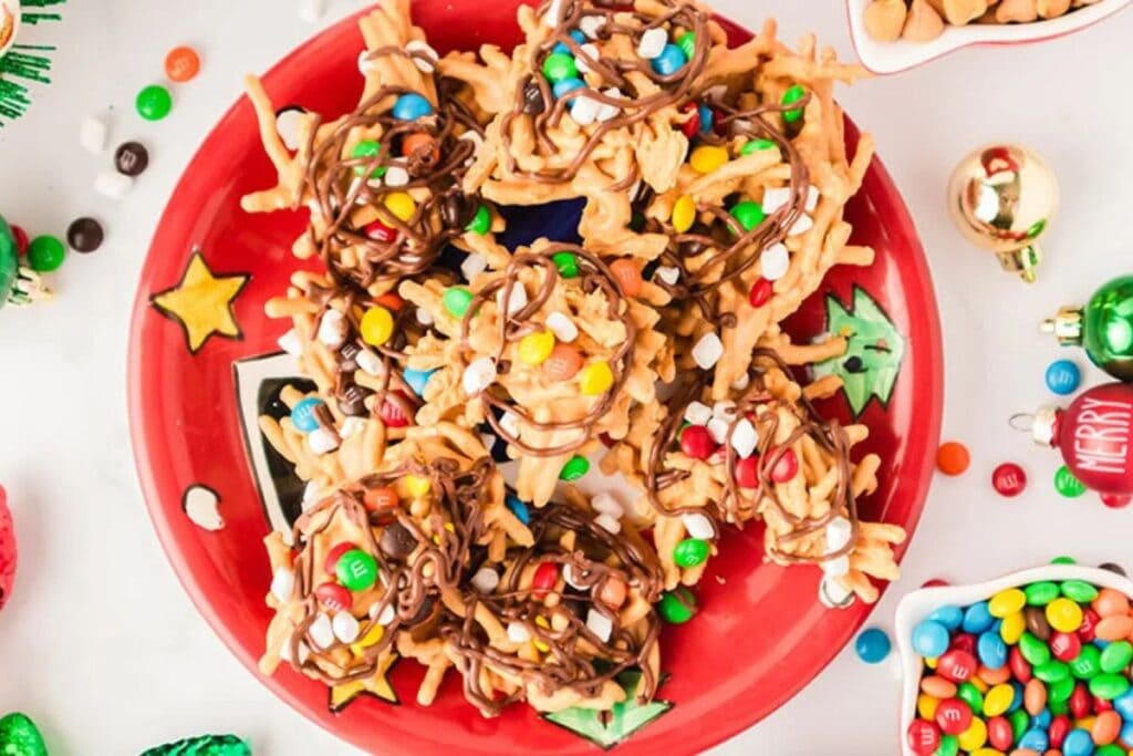 Haystack cookies with chow mein noodles, peanut butter, chocolate drizzle, and M&Ms on a red plate.