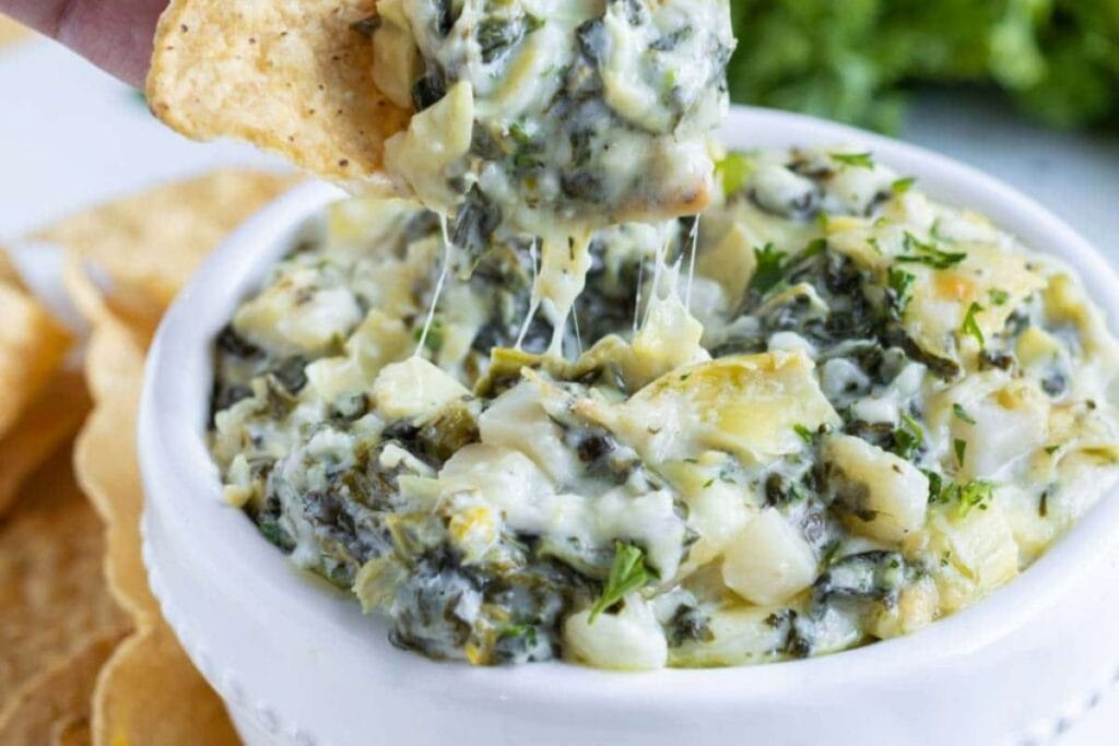 Tortilla chip scooping cheesy spinach artichoke dip from a white bowl.
