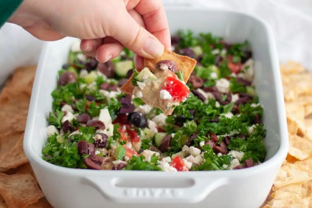 Hand dipping a pita chip into a layered Greek hummus dip.