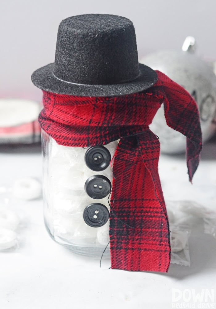 Glass jar decorated as a snowman with black buttons, plaid scarf, and felt top hat, filled with white candies on a light surface.
