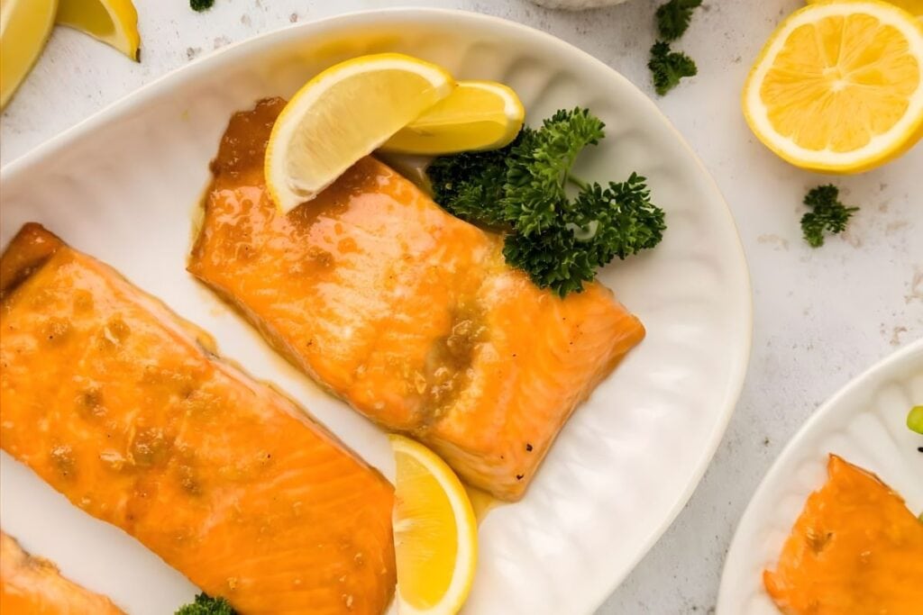 Two maple-glazed salmon fillets on a white plate with lemon and parsley.