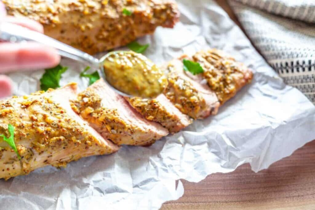Sliced pork tenderloin being drizzled with mustard sauce on parchment paper.