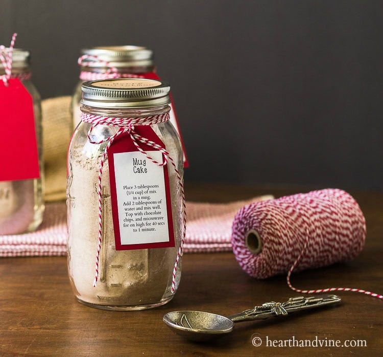 Glass jar labeled “Mug Cake” with powdered mix, tied with red-and-white twine and red tag, surrounded by decorated jars, twine spool, and spoon on wooden surface.