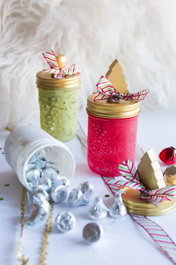 Three decorated mason jars in red, green, and white with gold lids and striped bows, one tipped to spill silver Hershey’s Kisses, surrounded by beads, sequins, and jingle bell on a fluffy white surface.