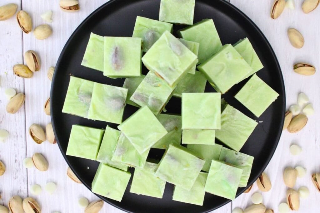 Green pistachio fudge squares on a black plate with pistachios around.