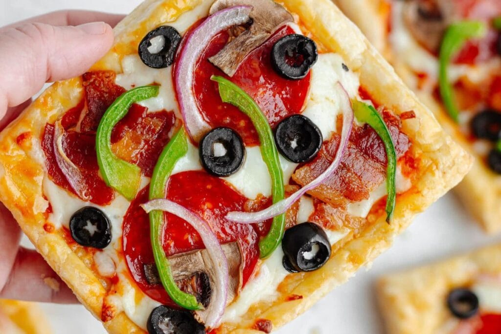 Hand holding a square slice of puff pastry pizza with toppings.