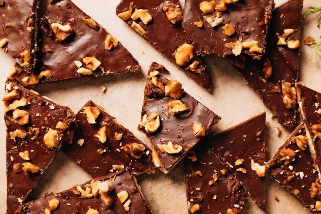 Chocolate-covered saltine cracker toffee pieces topped with nuts, arranged on a light brown surface.