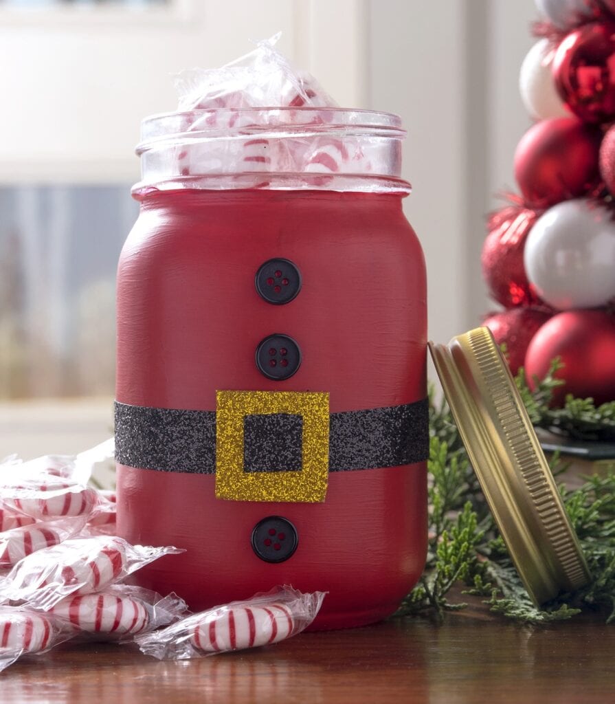 Red jar decorated like Santa with black belt and gold buckle, filled with peppermint candies and surrounded by festive ornaments on a wooden surface.