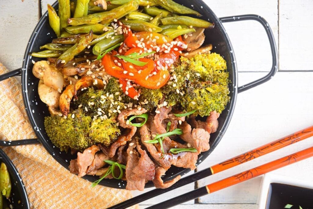 Beef and broccoli stir-fry in a black pan with green beans and red peppers.