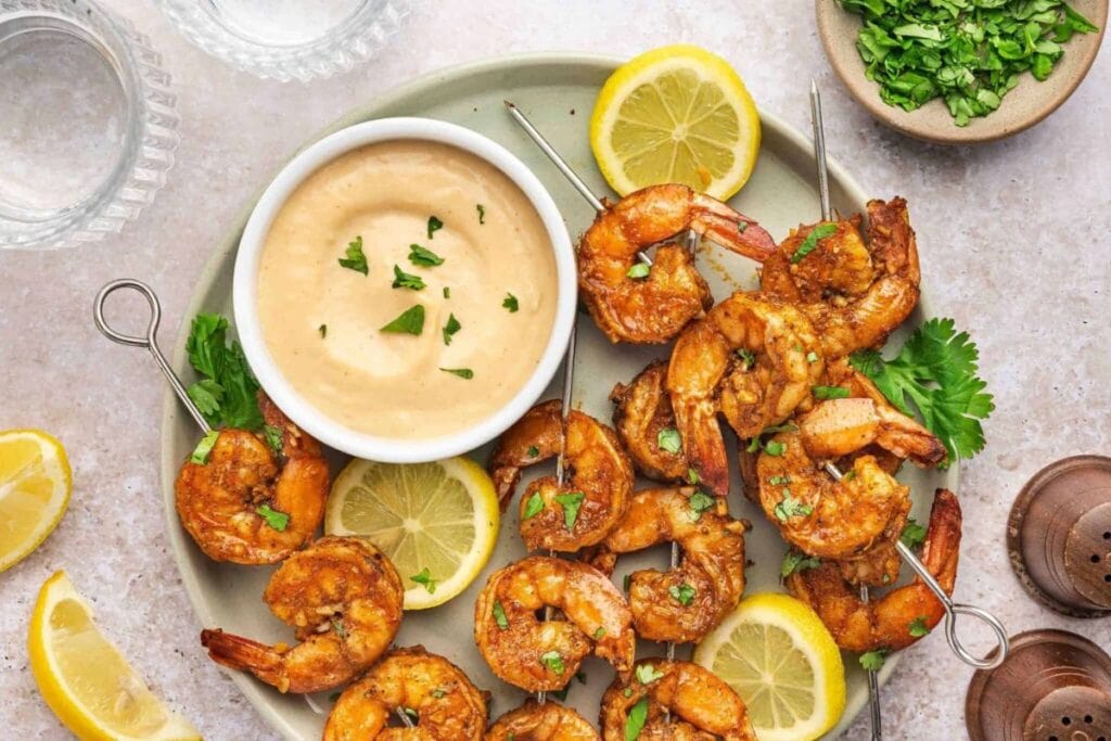 Shrimp skewers on a plate with dipping sauce and lemon wedges.