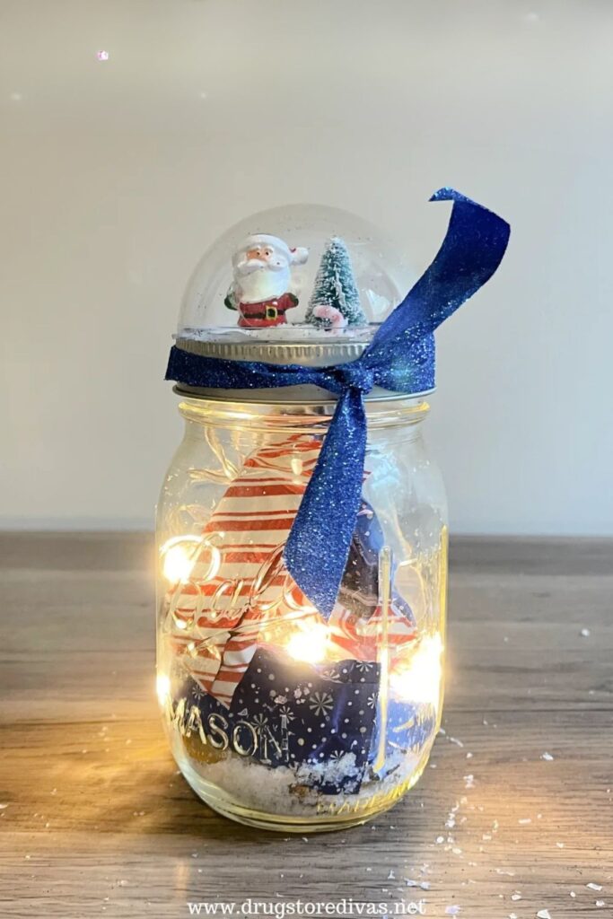 Clear mason jar with miniature holiday scene, filled with artificial snow, striped cone, and string lights, topped with a snow globe lid featuring Santa and a Christmas tree, tied with a blue ribbon.