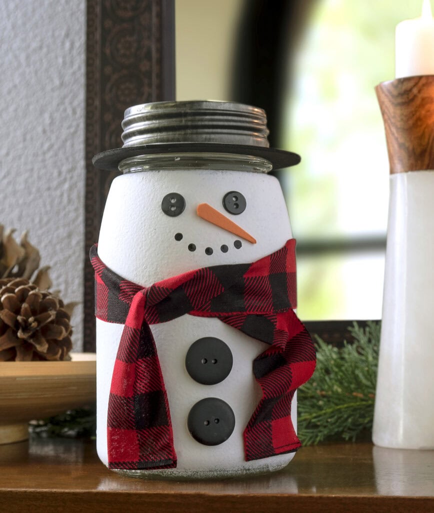 White-painted mason jar decorated as a snowman with button eyes, carrot nose, plaid scarf, and metal lid hat, placed on wood with pinecone, greenery, and candle nearby.
