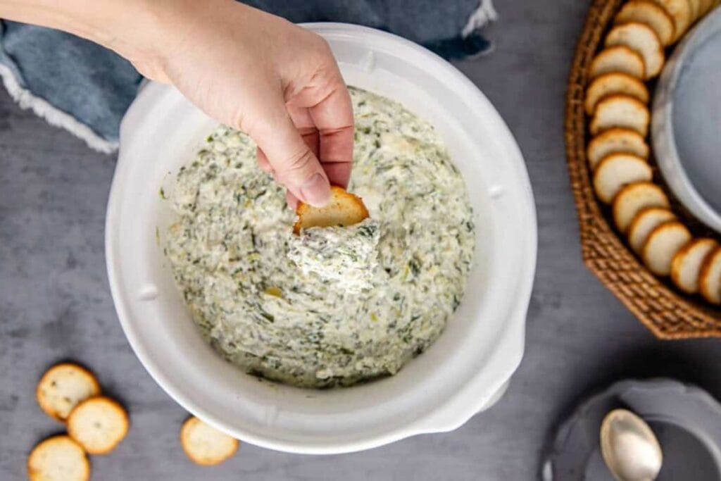 A cracker being dipped into a bowl of spinach and artichoke dip.