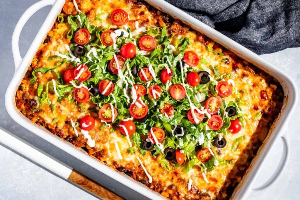 Taco casserole in a white dish, topped with lettuce, tomatoes, olives, and sour cream.