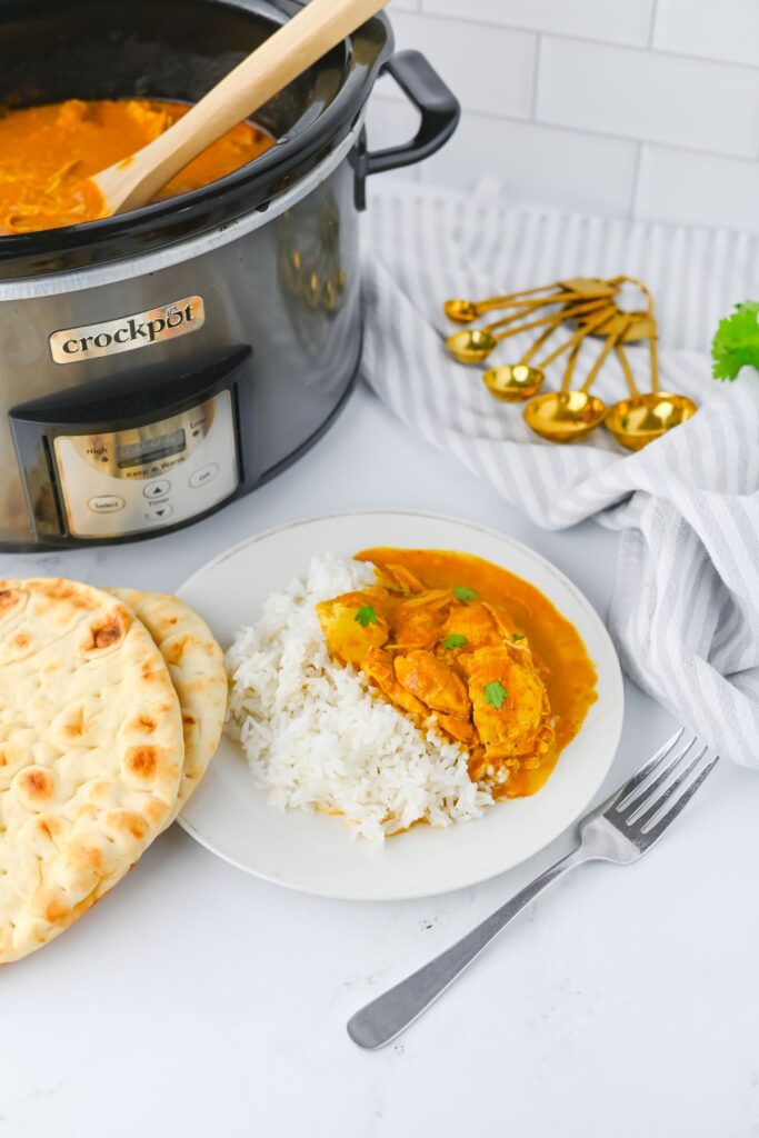 Butter chicken served over rice next to warm naan and a slow cooker.