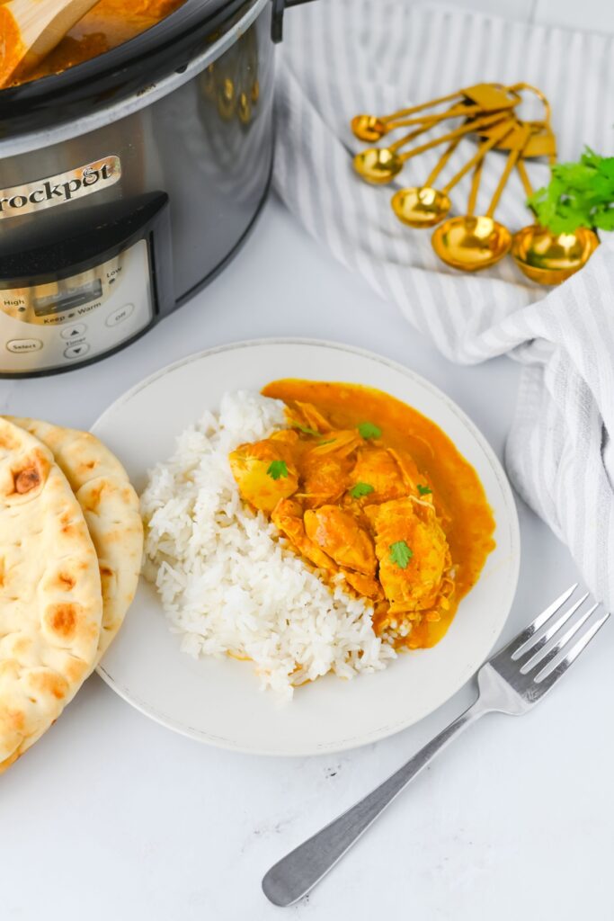 Butter chicken served over rice next to warm naan and a slow cooker.