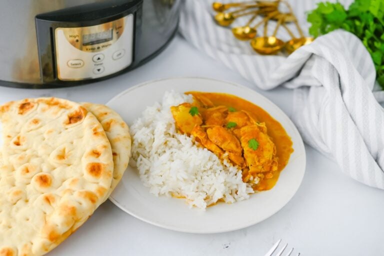 Butter chicken served over rice next to warm naan and a slow cooker.