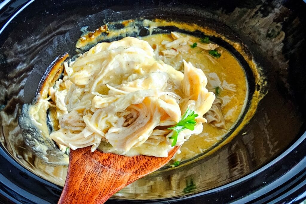 Shredded chicken and gravy scooped from a slow cooker on a wooden spoon.