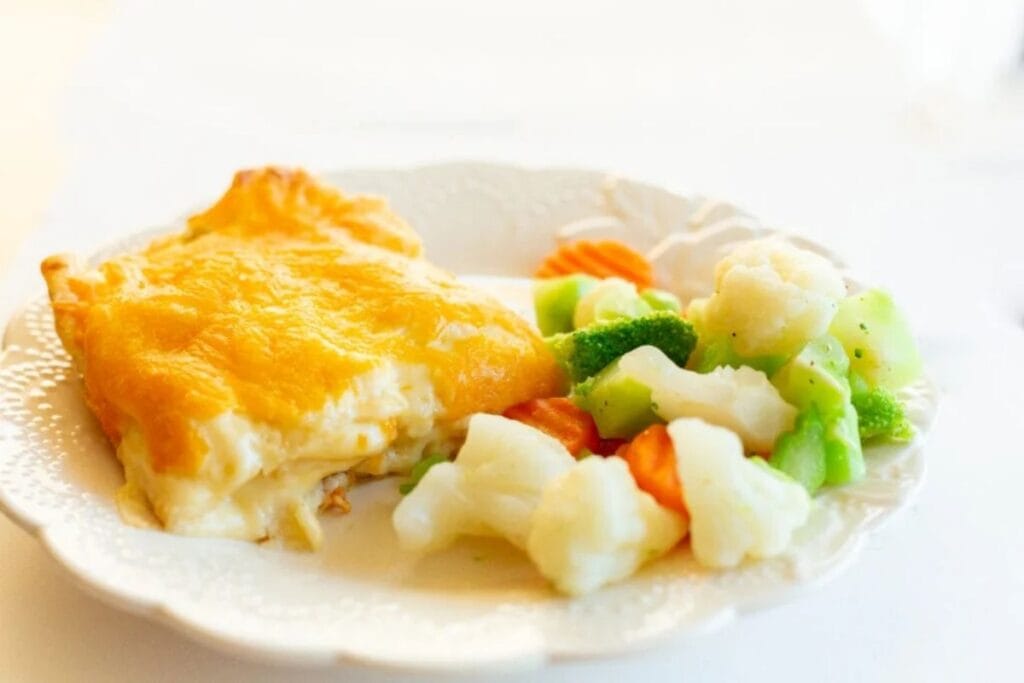 Slice of cheesy chicken enchilada casserole served with steamed broccoli, cauliflower, and carrots.