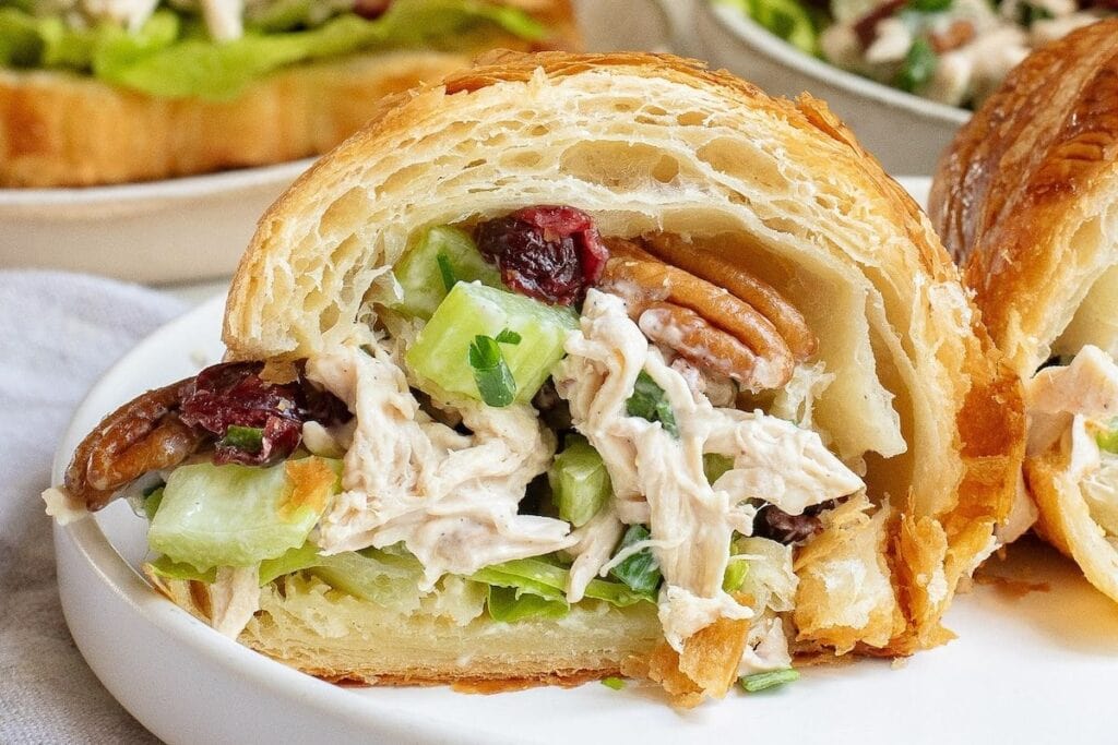 Croissant sandwich filled with chicken salad, celery, pecans, and cranberries on a white plate.