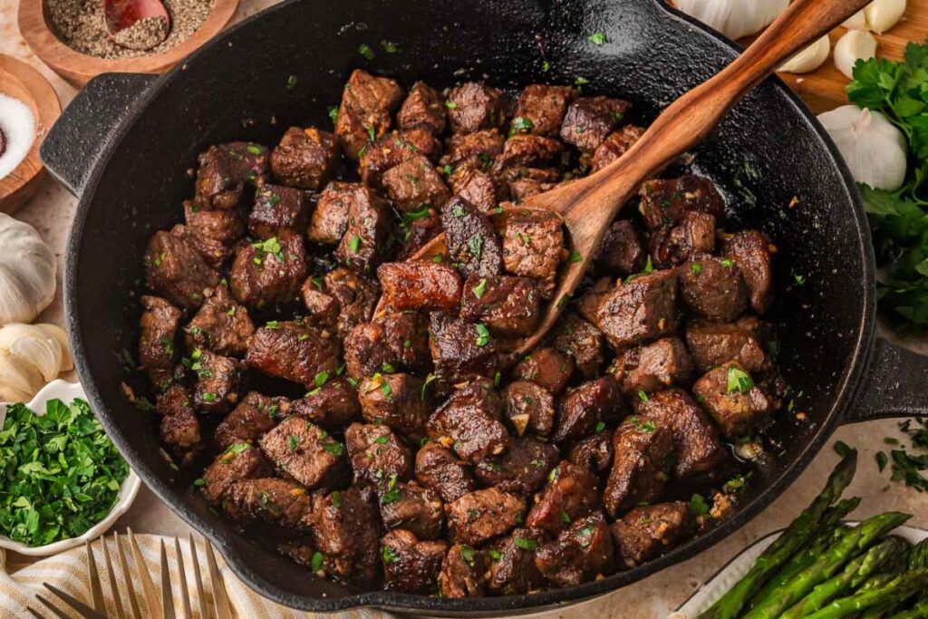 Seared steak bites in a cast-iron skillet with fresh parsley and a wooden spoon.