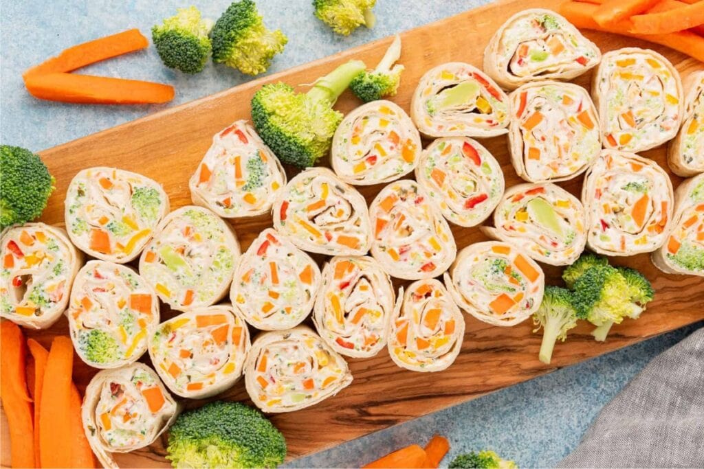 Veggie pinwheels on a wooden board with broccoli and carrots.