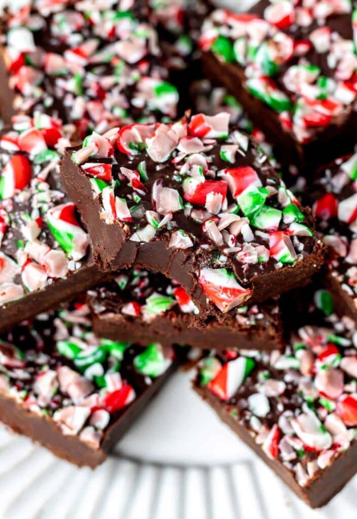 Chocolate fudge squares topped with crushed red, white, and green peppermint candy, stacked on a white plate for a holiday treat.