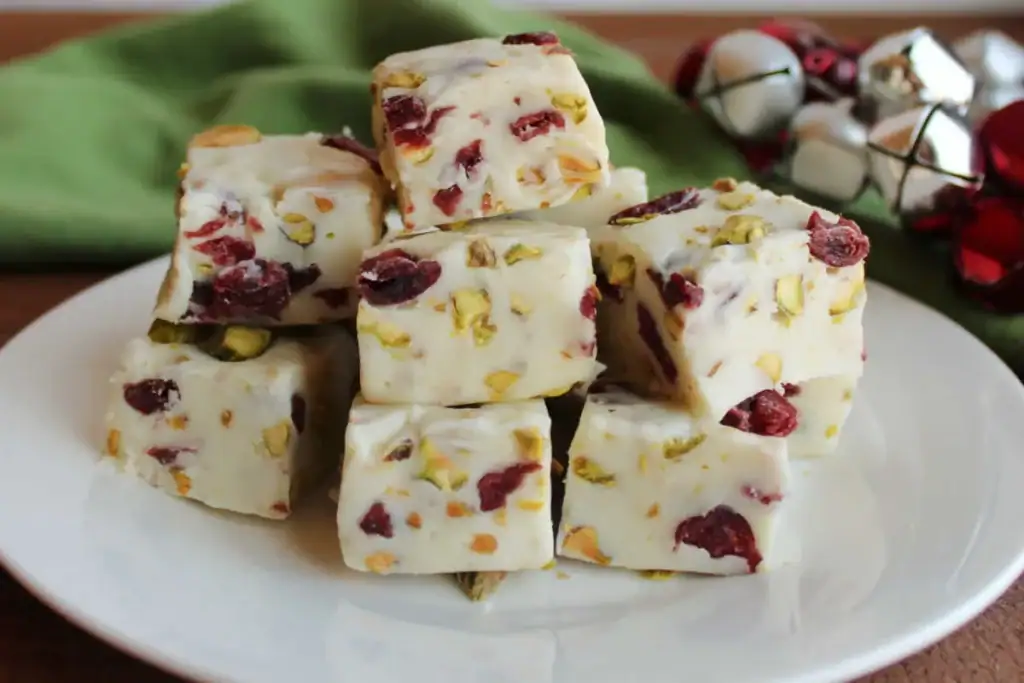 White fudge cubes with cranberries and pistachios, stacked on a plate with festive jingle bells and green cloth in the background.