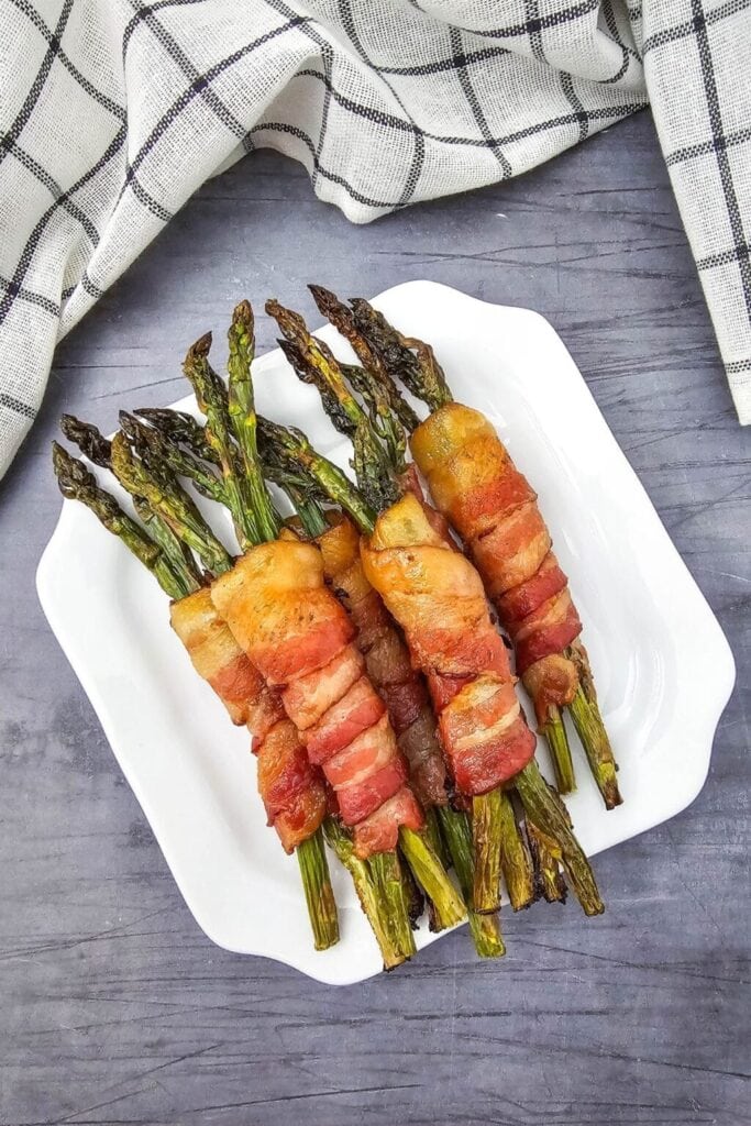 White plate of roasted asparagus bundles wrapped in golden bacon, arranged neatly on a gray surface with checkered cloth in background.