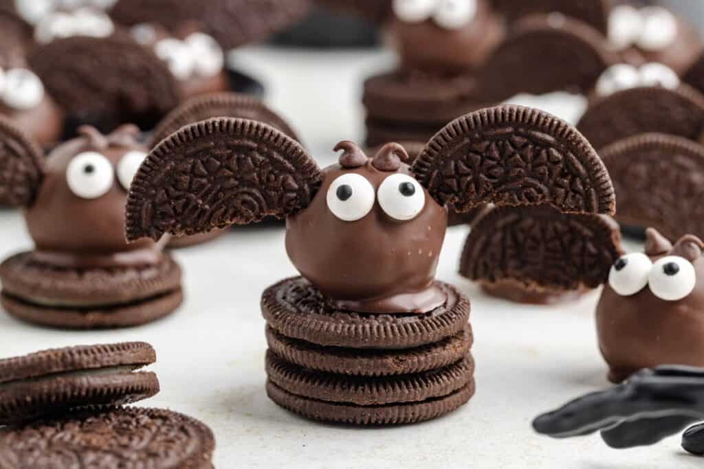 Chocolate Halloween treats decorated as bats with cookie ears, candy eyes, and chocolate-coated centers, displayed on stacked chocolate sandwich cookies.