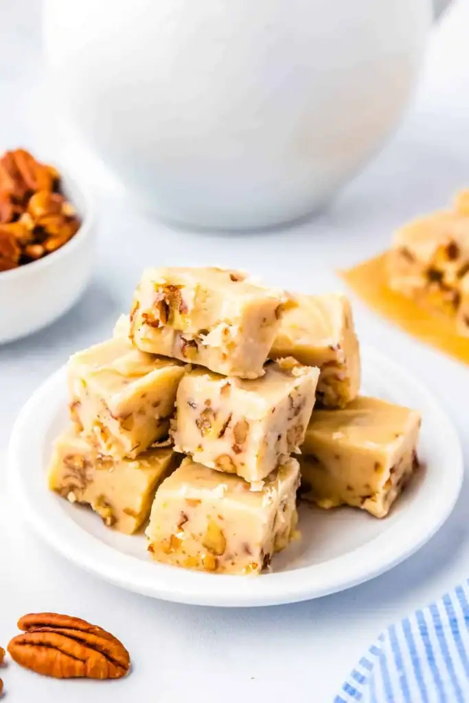 Light brown fudge cubes with pecan pieces, stacked on a plate beside a bowl of nuts and a white teapot.
