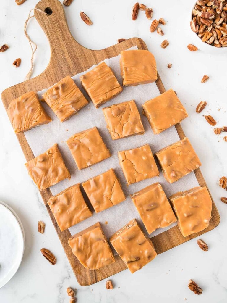 Caramel-colored fudge squares arranged on a wooden board with scattered pecans and a bowl of nuts nearby.