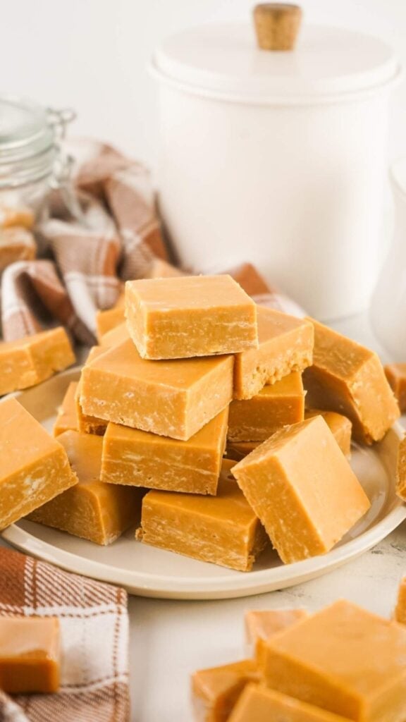 Caramel-colored fudge squares stacked on a plate, surrounded by rustic kitchen props including a checkered cloth, jar, and ceramic container.