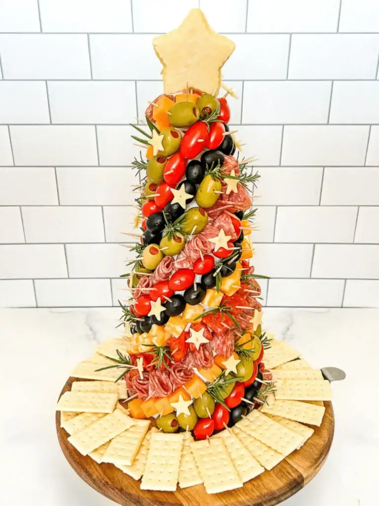 Christmas tree–shaped charcuterie board with olives, meats, cheeses, tomatoes, and rosemary sprigs, topped with a cheese star and surrounded by crackers on a wooden board.