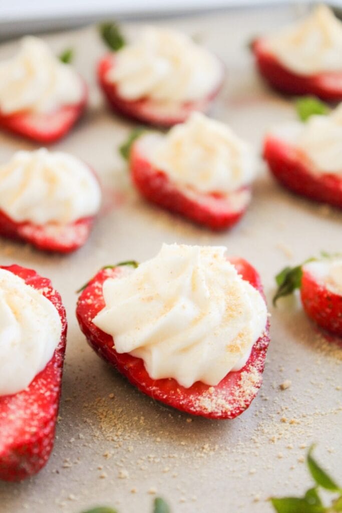 Halved strawberries topped with creamy cheesecake filling and graham crumbs, arranged on a light surface with green leaves attached.