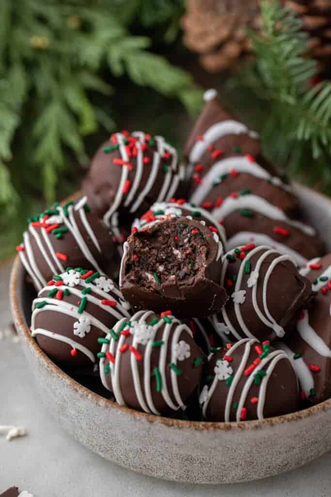 Chocolate-coated Christmas tree cake truffles decorated with white chocolate drizzle and festive red, green, and white sprinkles, with one truffle cut open to reveal the chocolate cake filling.