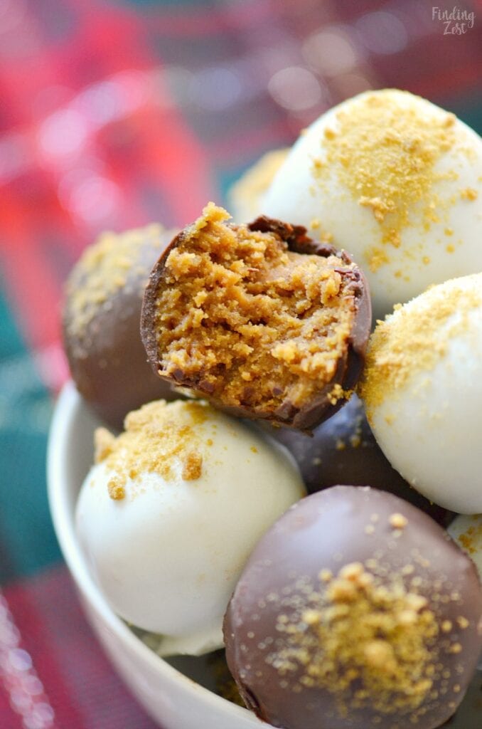 Close-up of a bowl of chocolate truffles coated in white and milk chocolate with crumb topping, one bitten to reveal a golden interior, set against a festive red and green plaid background.