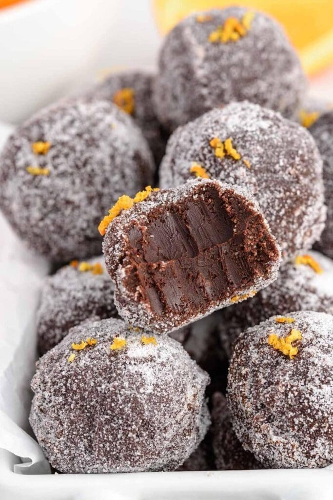 Close-up of chocolate truffles coated in sugar, garnished with orange zest, with one truffle cut open to reveal a smooth chocolate interior in a white dish.