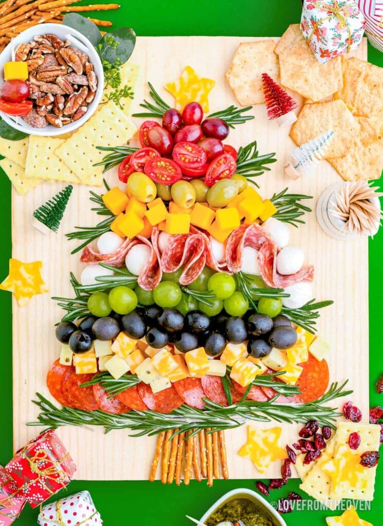 Christmas tree–shaped charcuterie board layered with grapes, cheeses, meats, olives, and rosemary sprigs, topped with a cheese star and pretzel trunk, surrounded by crackers, nuts, and holiday decorations.